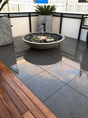 Circular concrete water fountain on wet bluestone paving with timber decking and white planters, St Kilda rooftop
