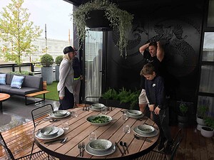 Round timber dining table set for outdoor meal on St Kilda rooftop terrace with people preparing food nearby