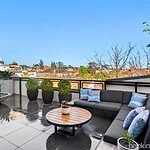 Grey sectional sofa with blue cushions on rooftop terrace overlooking St Kilda cityscape with round timber coffee table