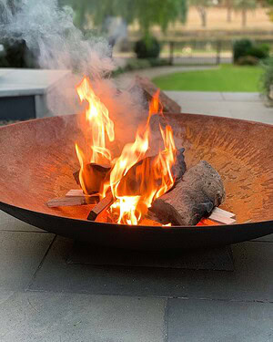 Iron fire bowl with flames burning wood on paved outdoor area with lawn and house in background